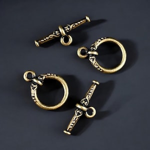 May include: Four antique brass toggle clasps. Two are circular with decorative detailing, and two are bar-shaped with a loop. The clasps are arranged on a dark gray surface. These clasps are ideal for jewelry making and other craft projects.