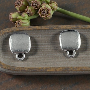 May include: Two silver-toned metal charms with a square shape and rounded corners. Each charm features a smooth, slightly textured surface and a small circular loop at the bottom for attachment. The metal has a matte, brushed finish.