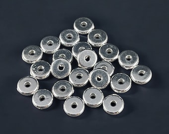 May include: A collection of shiny, silver-colored metal beads with a flat, circular shape and a hole in the center. The beads are scattered on a dark gray surface, creating a visually appealing arrangement. These beads are ideal for jewelry making.
