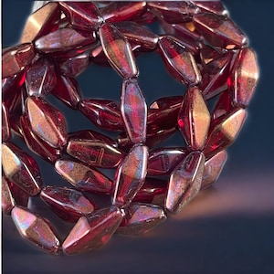 May include: Close-up of a strand of red, iridescent beads. The beads are elongated, with a faceted, oblong shape. The beads have a metallic sheen, reflecting light with hints of gold and purple. The beads are strung together, forming a circular shape.