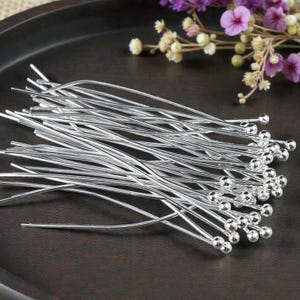 May include: A collection of silver-colored head pins with small, round ball ends. The pins are arranged in a pile on a dark brown surface, with some floral accents in the background. These pins are likely used for jewelry making or crafting.