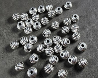 May include: A collection of silver-colored, spiral-grooved beads. Each bead has a cylindrical shape with a hole running through the center. The beads are scattered on a dark gray surface, creating a textured appearance.