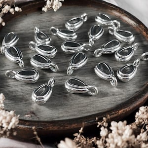 May include: A close-up of a group of silver teardrop-shaped pendant bails. The bails are arranged on a wooden surface.