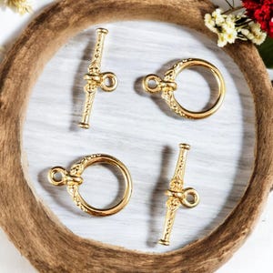 May include: Four gold-colored toggle clasps for jewelry making. Two clasps are circular, and two are bar-shaped. The clasps have an ornate design and are arranged on a white surface, surrounded by a wooden frame.