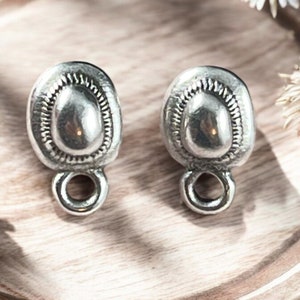 May include: Two silver-toned oval-shaped earring components with a textured border and a loop at the bottom. The earrings have a slightly domed center and are displayed on a wooden surface.