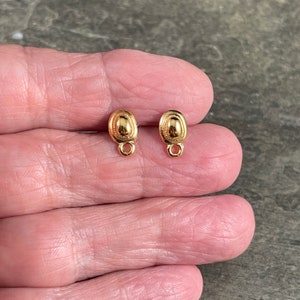 May include: Two small, gold-colored oval earring components with a textured design and a loop at the bottom. The earrings are displayed on a hand, showcasing their size and detail. The components are likely for jewelry making.
