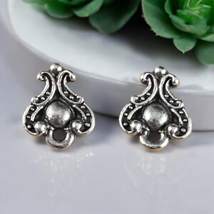 May include: A pair of silver-toned metal jewelry components featuring an ornate, symmetrical shape with a central circular dome, curved scrollwork details, and a small circular opening at the base, finished with darkened recessed areas.