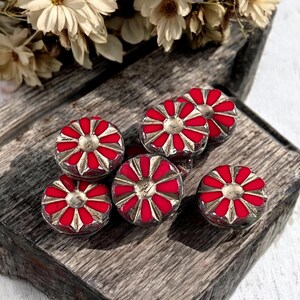 May include: Six red and silver-toned decorative beads with a floral design are arranged on a weathered wooden surface. The beads have a central silver-toned circle surrounded by red petals. Dried flowers are in the background.