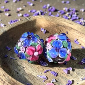 May include: Two spherical glass beads featuring intricate floral patterns in shades of blue, pink, and green. The beads have a smooth, glossy finish with raised petal details, showcasing a multi-colored design that covers the entire surface of each round bead.