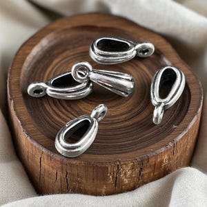 May include: A collection of four silver-toned metal jewellery bails, each featuring a smooth, teardrop-shaped loop with a small circular attachment point at the top, designed for securing pendants to necklaces or other jewellery components.