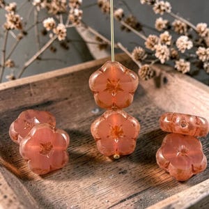 May include: Five translucent, peach-colored glass beads shaped like five-petaled flowers. Two beads are stacked vertically on a thin gold-toned metal pin, while three individual beads are arranged around them on a wooden surface.