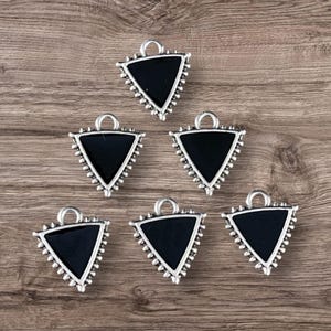 May include: Six silver-toned triangle pendants with a black enamel center and a beaded edge. Each pendant has a small loop at the top for attaching to a chain or other jewelry component. The pendants are displayed on a wooden surface.