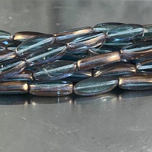 May include: Close-up of a strand of iridescent glass beads. The beads are oval-shaped with a longitudinal stripe, displaying shades of blue, green, and bronze. The beads are strung together, creating a textured surface.