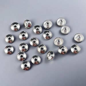 May include: A collection of 18 silver-toned metal beads with a flat, round shape and a raised center. The beads have a smooth, shiny finish.