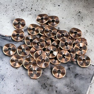 May include: A pile of 25 copper-colored metal beads with a raised design. The beads are round with a hole in the center.