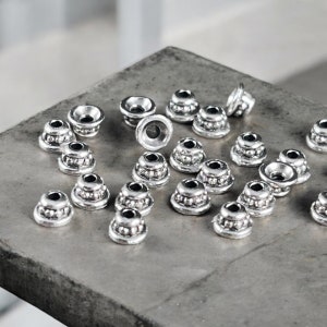 May include: A collection of small, silver-toned metal beads with a tiered, conical shape. Each bead features a central hole and a textured, beaded base detail. The metallic finish has a slightly oxidized appearance, highlighting the intricate circular patterns on the exterior surface of these craft components.