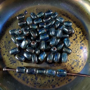 May include: A collection of small, barrel-shaped beads with a glossy, dark blue and black iridescent finish. The beads feature a central hole and are shown both in a loose pile and threaded in a single row on a thin, dark metal wire.