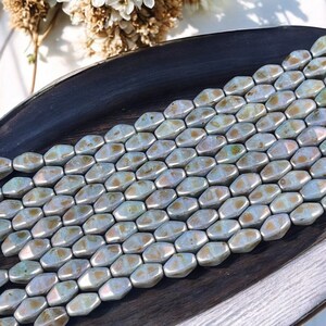 Pode incluir: Uma cole&ccedil;&atilde;o de contas hexagonais iridescentes em tons de prata, dourado e azul. As contas est&atilde;o dispostas em fileiras ordenadas, refletindo a luz e criando um efeito brilhante. As contas s&atilde;o exibidas em uma superf&iacute;cie de madeira escura.
