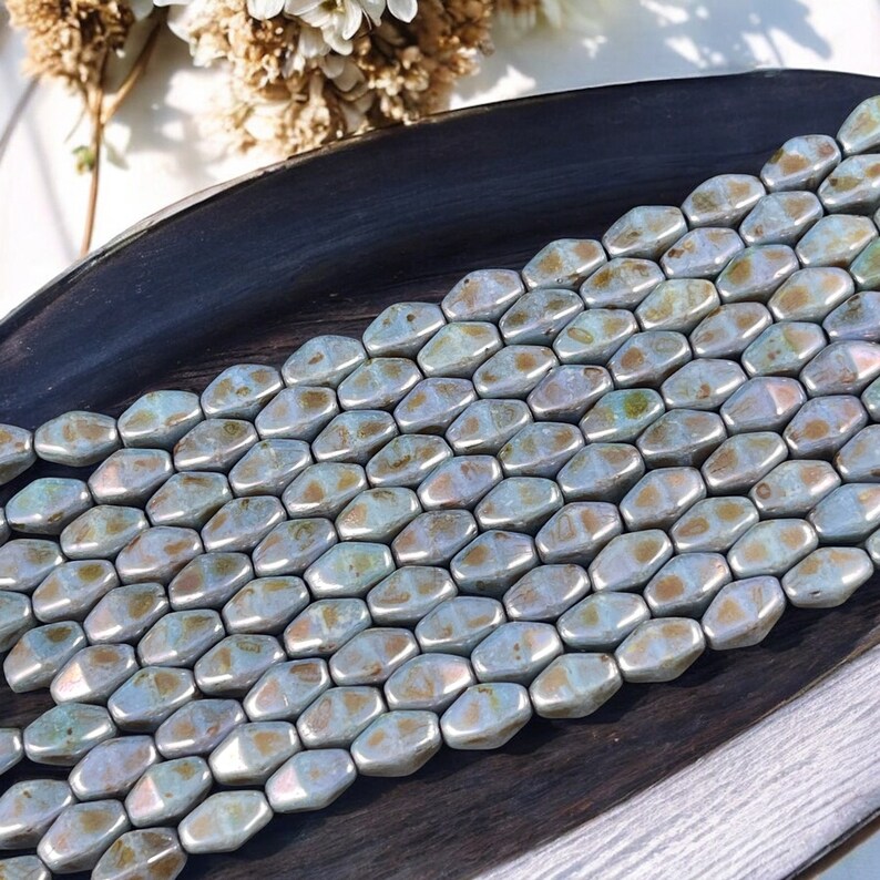 Pode incluir: Uma cole&ccedil;&atilde;o de contas hexagonais iridescentes em tons de prata, dourado e azul. As contas est&atilde;o dispostas em fileiras ordenadas, refletindo a luz e criando um efeito brilhante. As contas s&atilde;o exibidas em uma superf&iacute;cie de madeira escura.