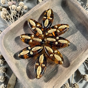 May include: A collection of oval-shaped, amber-colored beads arranged in a flower-like pattern. Each bead has a dark brown border and a gold-colored, textured design in the center. The beads are displayed on a wooden tray.