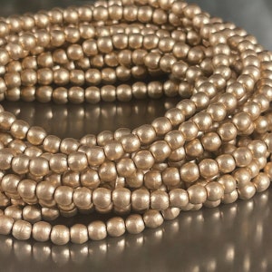 May include: Close-up of a coiled strand of metallic gold beads. The beads are small, round, and have a matte finish. The beads are strung together, creating a continuous line. The overall effect is one of elegance and simplicity, suitable for jewelry making.