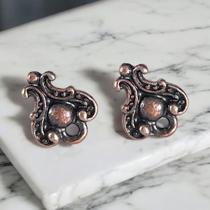 May include: A pair of ornate copper-toned stud earrings. Each earring features a central circular element surrounded by swirling, decorative details and small, raised spheres. The earrings are displayed on a white marble surface.