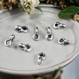 May include: Eight silver-toned metal jump rings with a small loop at the top. The jump rings are arranged on a white ceramic surface.