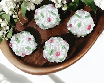 May include: Four white floral-shaped objects with pink centers and green accents are arranged in a wooden bowl. The bowl is brown and has a curved shape. White flowers and green leaves are in the background.