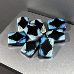 May include: A collection of rectangular glass beads featuring a central black diamond-shaped inlay surrounded by a metallic, iridescent blue and silver finish. Each bead has a smooth, polished surface with distinct edges and a reflective sheen.