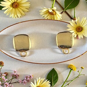 May include: Two gold-toned metal jewelry components, each featuring a smooth, square-shaped plate with rounded corners and a small circular loop attached at the bottom, resting on an oval-shaped white dish with a speckled texture.