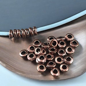May include: Copper-colored metal beads with a hammered texture. Some beads are strung on a light blue cord, while others are scattered on a copper-toned tray. The beads have a hole in the center.
