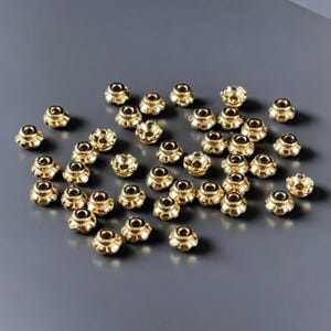 May include: A collection of small, gold-colored beads with a flower-like design. The beads are scattered on a gray surface, creating a visually appealing arrangement. These decorative beads are ideal for jewelry making and craft projects.