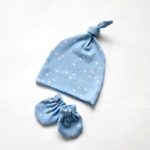 May include: A light blue baby hat with a white cross pattern and matching mittens. The hat has a knot on top.