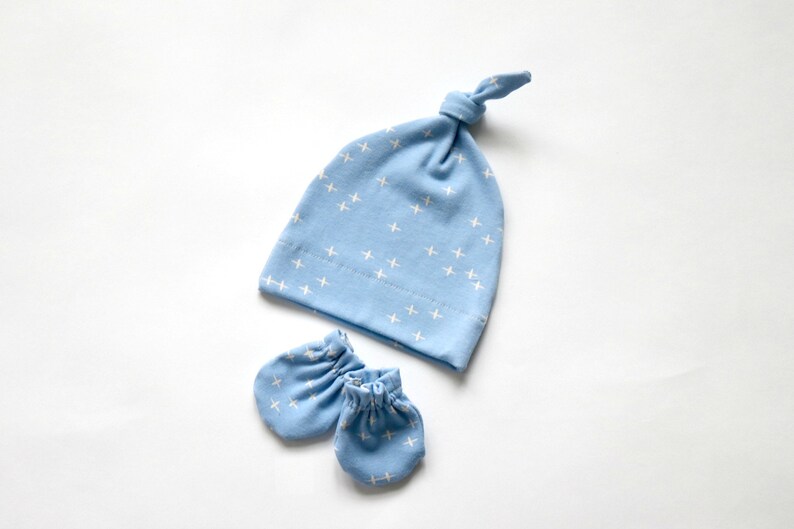 May include: A light blue baby hat with a white cross pattern and matching mittens. The hat has a knot on top.