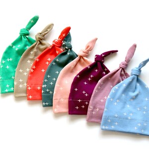 May include: Seven baby hats in different colors with a white plus sign pattern. The colors are teal, beige, coral, dark green, pink, purple, and light blue.