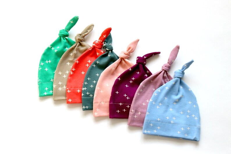 May include: Seven baby hats in different colors with a white plus sign pattern. The colors are teal, beige, coral, dark green, pink, purple, and light blue.