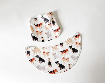 May include: Two white fabric bibs with a pattern of colourful dogs on a white background. The bibs are both in a triangular shape, with one being a standard triangle and the other being a half-circle.