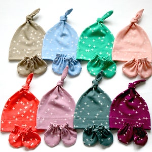 May include: Set of eight baby hats and matching mittens in various colors with a white plus sign pattern. The hats are tied at the top and the mittens are fingerless.
