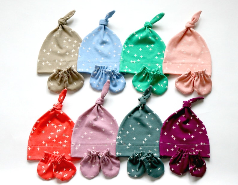 May include: Set of eight baby hats and matching mittens in various colors with a white plus sign pattern. The hats are tied at the top and the mittens are fingerless.