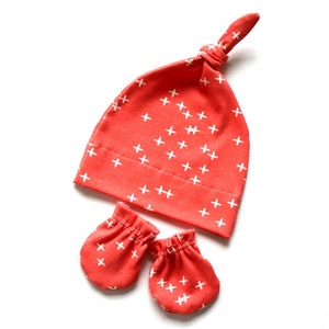 May include: A set of baby accessories in a coral color with a white plus sign pattern. The set includes a knotted hat and a pair of mittens.