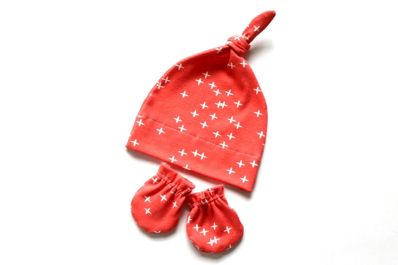 May include: A set of baby accessories in a coral color with a white plus sign pattern. The set includes a knotted hat and a pair of mittens.