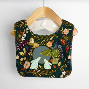 May include: A dark green baby bib with a floral pattern featuring white, pink, yellow, and red flowers, leaves, and butterflies. Two white rabbits are looking at each other in the centre of the bib.