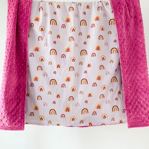 May include: A soft, pink and gray baby blanket with a rainbow pattern. The blanket is reversible, with a solid pink side and a side with a pattern of colorful rainbows on a gray background.