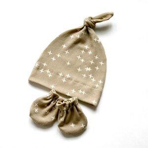 May include: A tan baby hat and matching mittens with a white plus sign pattern.