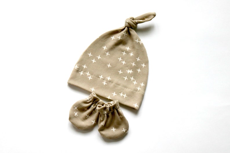 May include: A tan baby hat and matching mittens with a white plus sign pattern.