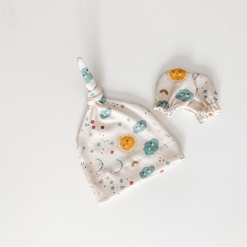 May include: A set of baby accessories featuring a cream-coloured knotted hat and matching mittens. The hat and mittens have a whimsical print of suns, clouds, and rainbows in shades of yellow, blue, and red.