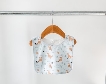 May include: A light blue baby bib with a woodland animal print featuring foxes, owls, and leaves. The bib has a white trim and a snap closure.