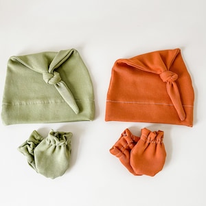 May include: Two sets of newborn baby accessories. One set is green and the other is orange. Each set includes a knotted hat and a pair of mittens.