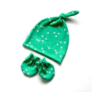 May include: A set of baby accessories in a green and white color scheme. The set includes a hat with a knotted top and a pair of mittens. Both the hat and mittens have a white cross pattern on a green background.