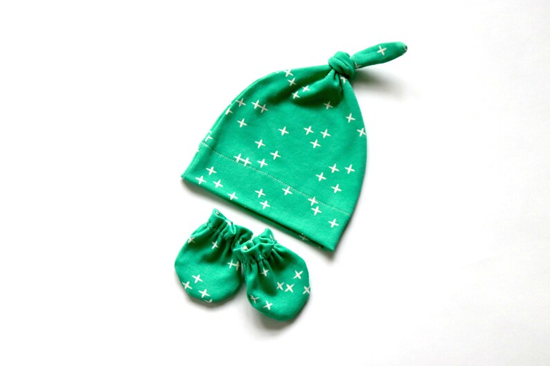 May include: A set of baby accessories in a green and white color scheme. The set includes a hat with a knotted top and a pair of mittens. Both the hat and mittens have a white cross pattern on a green background.